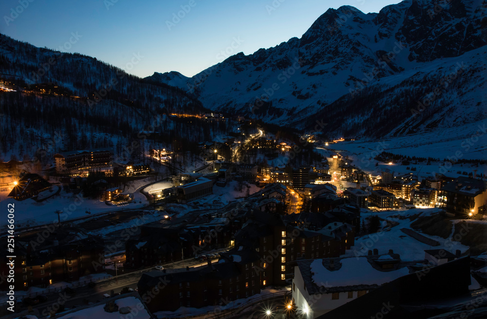 European mountains landscape, Alps, Italy, mountain skiing resort, picturesque view with deep blue sky, high rock peaks on horizon, town village houses lights at night photo. Tourism, travel, resort