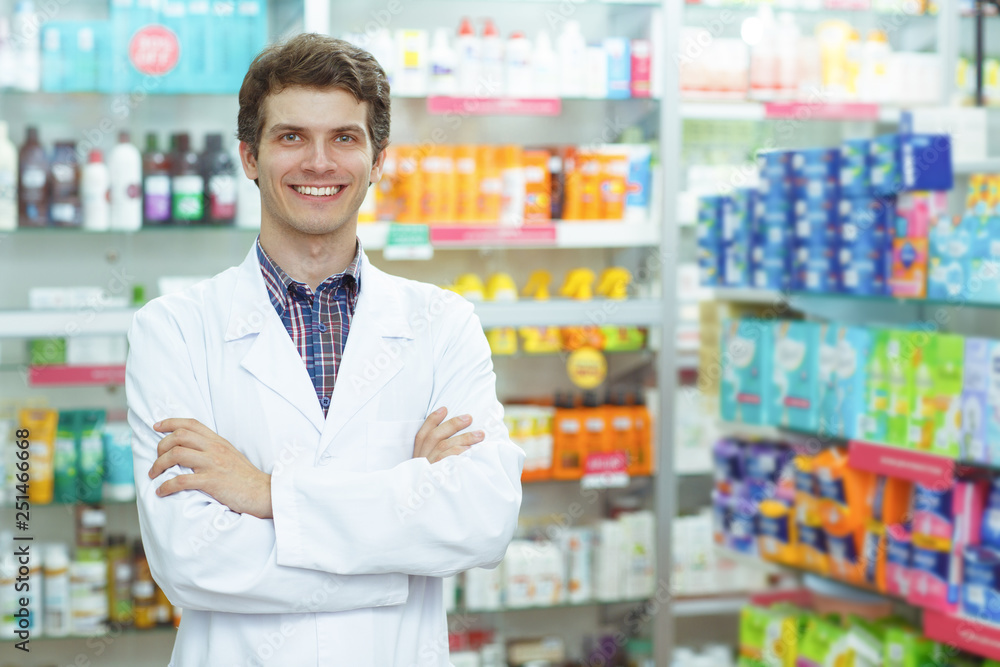 Front view of good looking pharmacist standing in drugstore. Handsome consultant smiling, looking at camera and posing. Background of shelves with medications.
