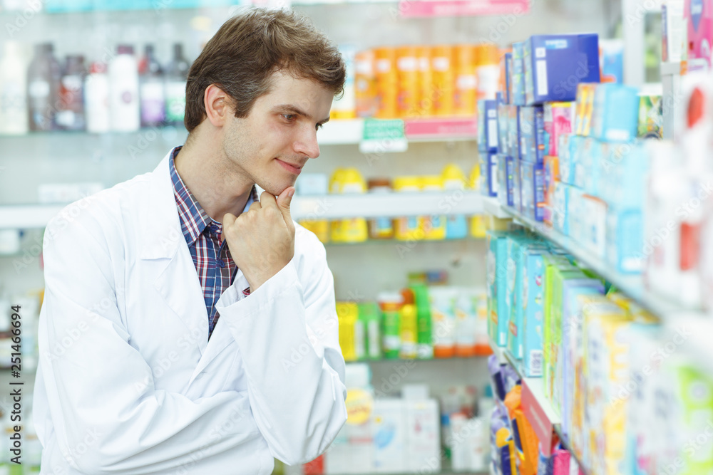 Serious male pharmacist holding hand near chin and looking at shelf with lots of medications. Young drugstore consultant wearing special white coat while working. Serious male pharmacist holding hand near chin and looking at shelf with lots of medications. Young drugstore consultant wearing special white coat while working.