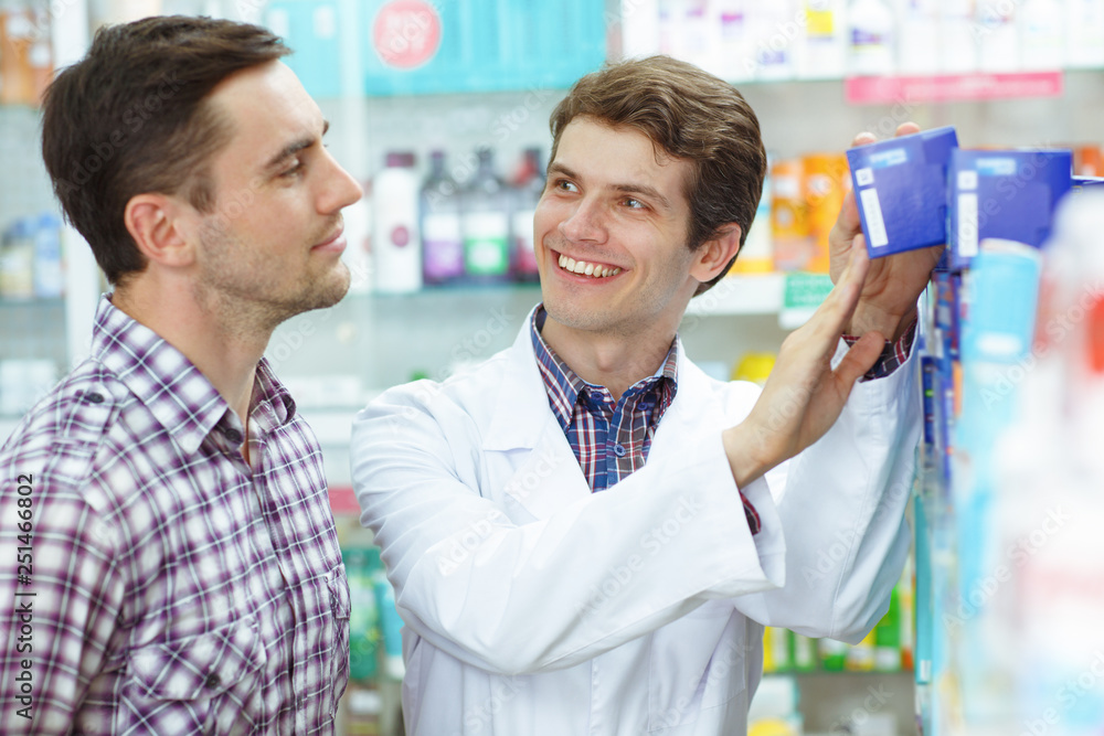 Pharmacist smiling, looking at customer and taking blue pack from shelf. Handsome man in checked shirt standing, looking at medicines and choosing. Men standing in drugstore.
