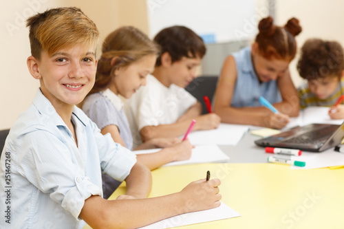 Stylish student of modern school in process of learning new material with classmates on background. Smiling  schoolboy sitting at table, writing in copybook, looking at camera and posing.