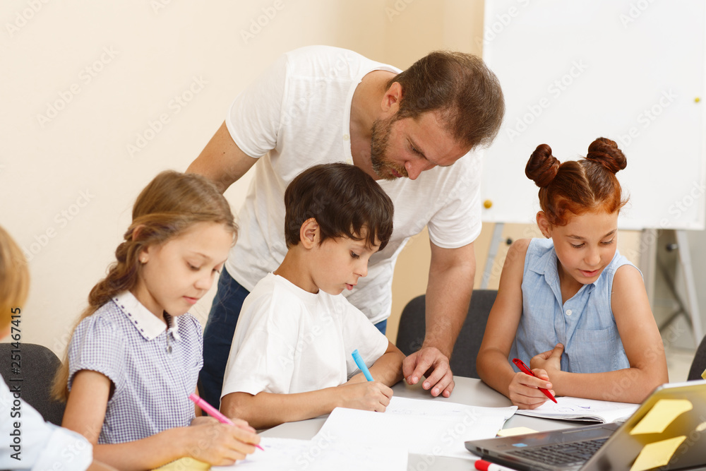 Serious male teacher in white shirt going around class, helping and checking works of pupils in classroom. Diligent little students of school sitting at table, writing and carefully doing tasks.