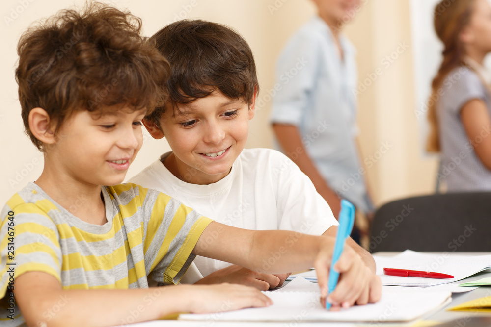 View from side of interested little pupils sitting together at table and writing in copybook. Smart classmates in process of learning new material in classroom. Concept of studying. View from side of interested little pupils sitting together at table and writing in copybook. Smart classmates in process of learning new material in classroom. Concept of studying.