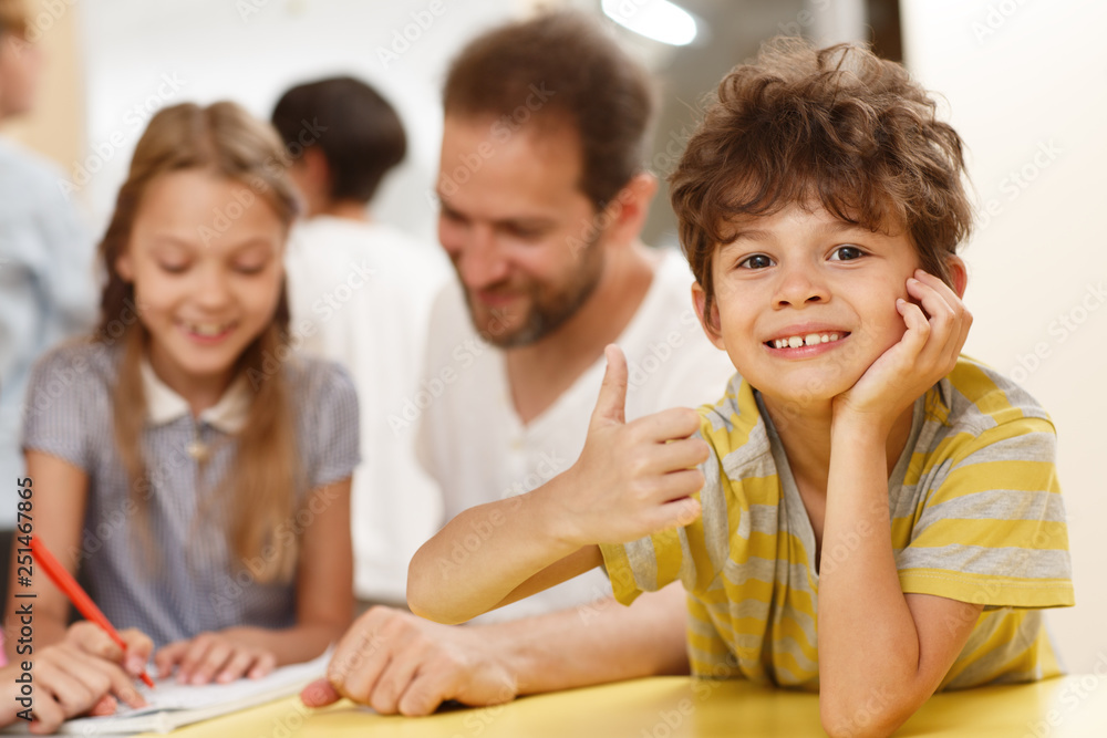 Cute pupil leaning on table, showing with hand sign OK, looking at camera and smiling in classroom. Little shoolboy enjoying interesting studying process. Concept of modern school. Cute pupil leaning on table, showing with hand sign OK, looking at camera and smiling in classroom. Little shoolboy enjoying interesting studying process. Concept of modern school.