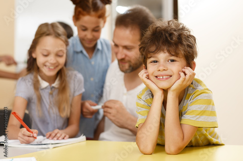 Portrait of shoolboy enjoying learning process in modern school. Little pupil smiling, looking at camera and leaning on table during lesson in classroom. Concept of Interesting education. © Zoriana