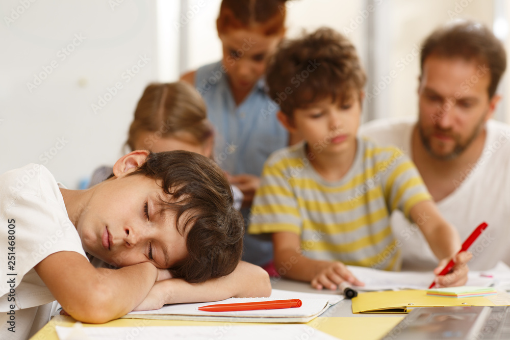 Lazy little student in white shirt sleeping during lesson in classroom. Exhausted with boring study boy leaning on table while classmates writing with teacher on background. Lazy little student in white shirt sleeping during lesson in classroom. Exhausted with boring study boy leaning on table while classmates writing with teacher on background.