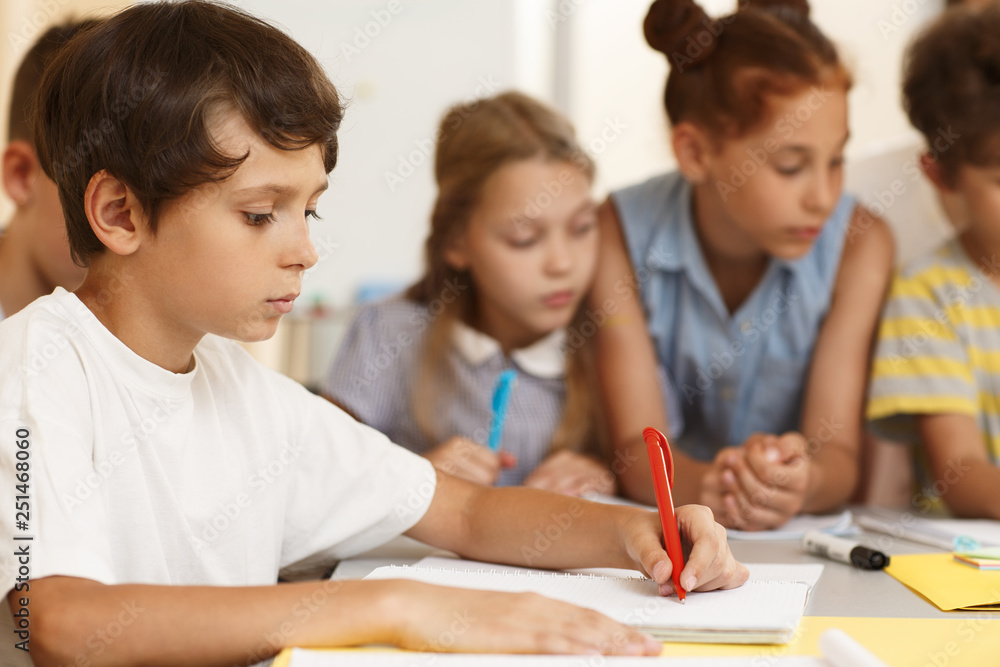Side view of serious little student sitting at table and writing essay in classroom. Hardworking pupil in white shirt doing test while classmates studying material in team. Concept of learning. Side view of serious little student sitting at table and writing essay in classroom. Hardworking pupil in white shirt doing test while classmates studying material in team. Concept of learning.