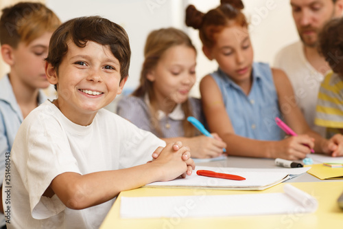 Smart classmates in process of learning new material in classroom. Little pupil sitting together at table and writing in copybook  one of group looking and smiling at camera.