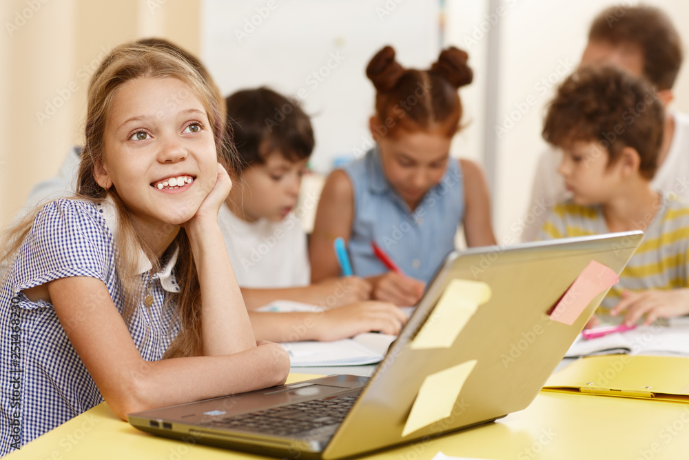 Portrait of smart pupil sitting at table, thinking ideas and looking away in classroom. Smiling little girl with classmates on background studying and using computer during lesson in school. Portrait of smart pupil sitting at table, thinking ideas and looking away in classroom. Smiling little girl with classmates on background studying and using computer during lesson in school.