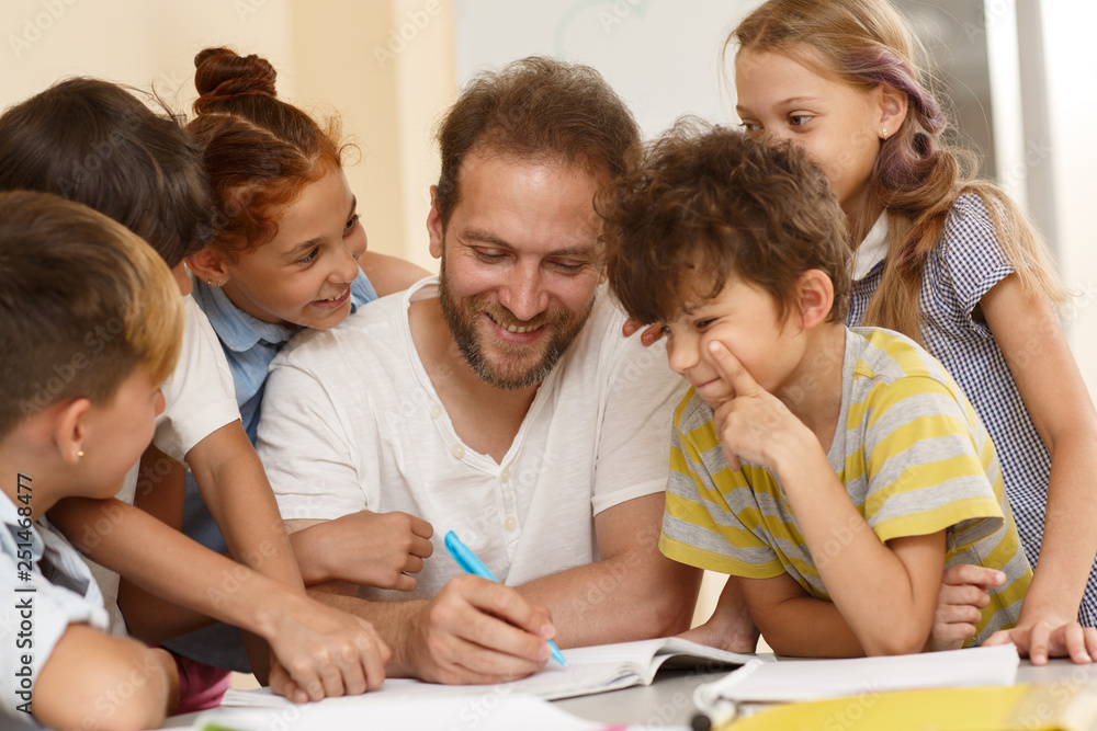 Positive male teacher in process of teaching little curious students in classroom. Smiling tutor sitting at table, writing and explaining material, smiling children standing around and listening. Positive male teacher in process of teaching little curious students in classroom. Smiling tutor sitting at table, writing and explaining material, smiling children standing around and listening.