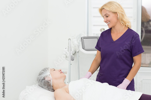 Professional dermatologist and client talking before hardware procedure in cosmetology office. Doctor in uniform and gloves looking at woman in disposable hat lying on couch. Apparatus on background.