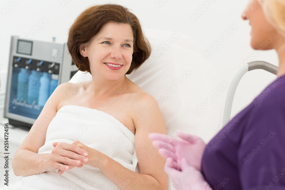 Obraz premium Woman having appointment with doctor cosmetologist. Female client lying on couch and talking with beautiful beautician in uniform and gloves. Concept of skin care and treatment.