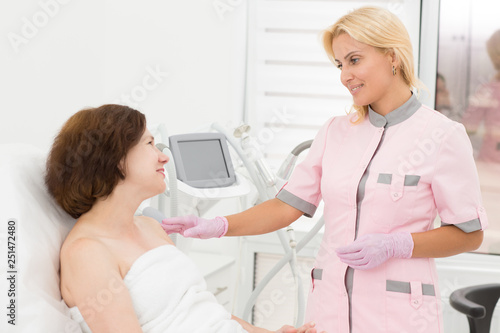 Professional doctor advising female client in cosmetology office. Beautician standing near woman lying on couch and talking with her. Consultation before hardware procedure. Skin care concept.