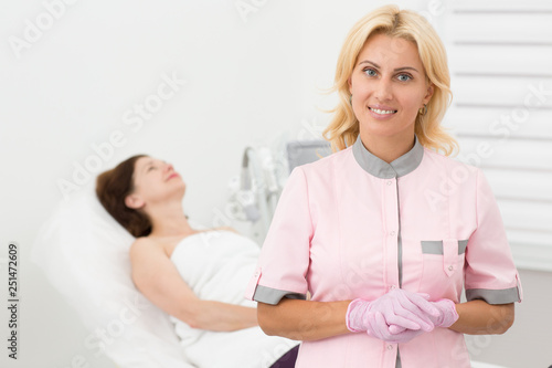 Portrait of female dermatologist during cosmetic anti aging procedure in cosmetology office. Pretty doctor posing  smiling and looking at camera. Satisfied patient lying on couch on background.