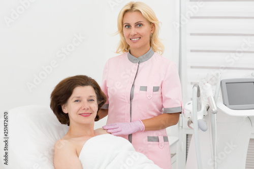 Doctor cosmetologist standing near beautiful client, looking at camera and posing. Satisfied woman looking wonderful after anti aging ultrasound procedure in cosmetology office. Skin care concept.