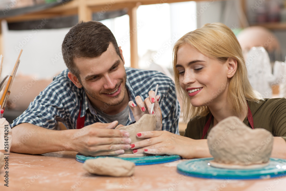 Side view of attractive craftswoman teaching handsome student art of making clay dishes in workshop. Lady sitting near man in checkered shirt and red apron and showing process of carving with knife. Side view of attractive craftswoman teaching handsome student art of making clay dishes in workshop. Lady sitting near man in checkered shirt and red apron and showing process of carving with knife.