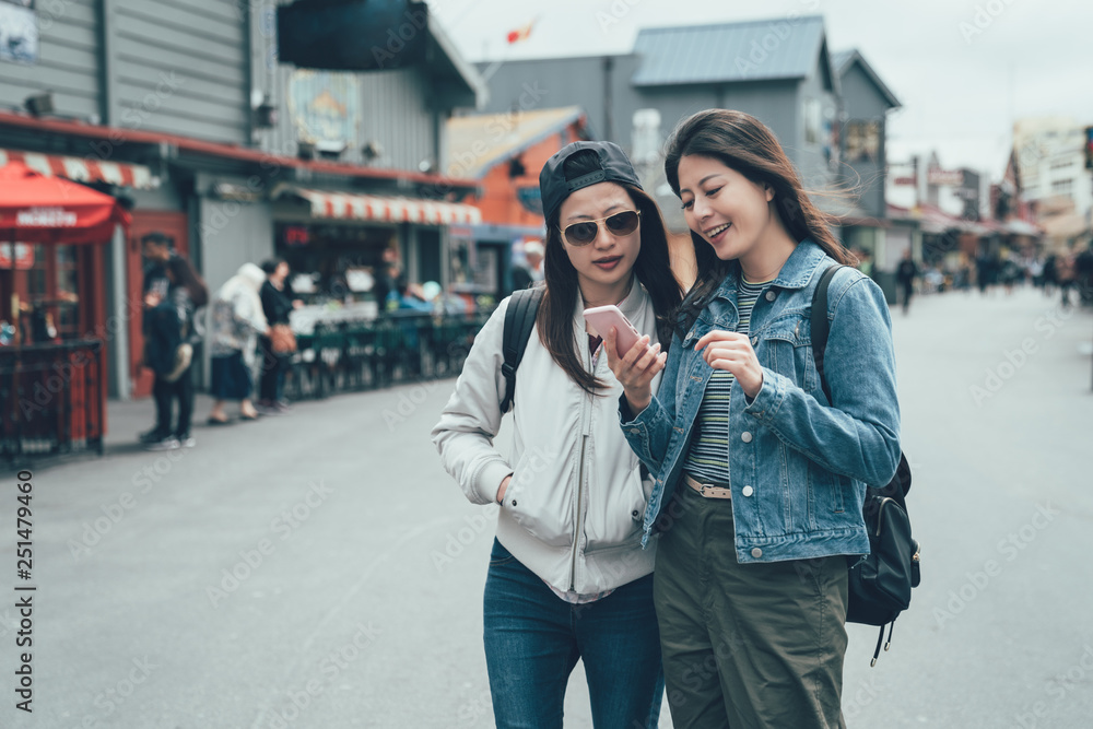 two young asian women friends of tourists looks at online map on cellphone standing outdoor farmer market in Old Fisherman's Wharf monterey. girl sisters smiling talk chat about mobile phone