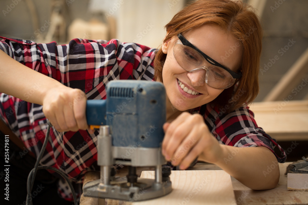 Female carpenter working with grinding machine in joiner's shop. Beautiful and positive woman with ginger hair smiling and grinding lumber. Professional craftswoman wearing safety glasses. Female carpenter working with grinding machine in joiner's shop. Beautiful and positive woman with ginger hair smiling and grinding lumber. Professional craftswoman wearing safety glasses.