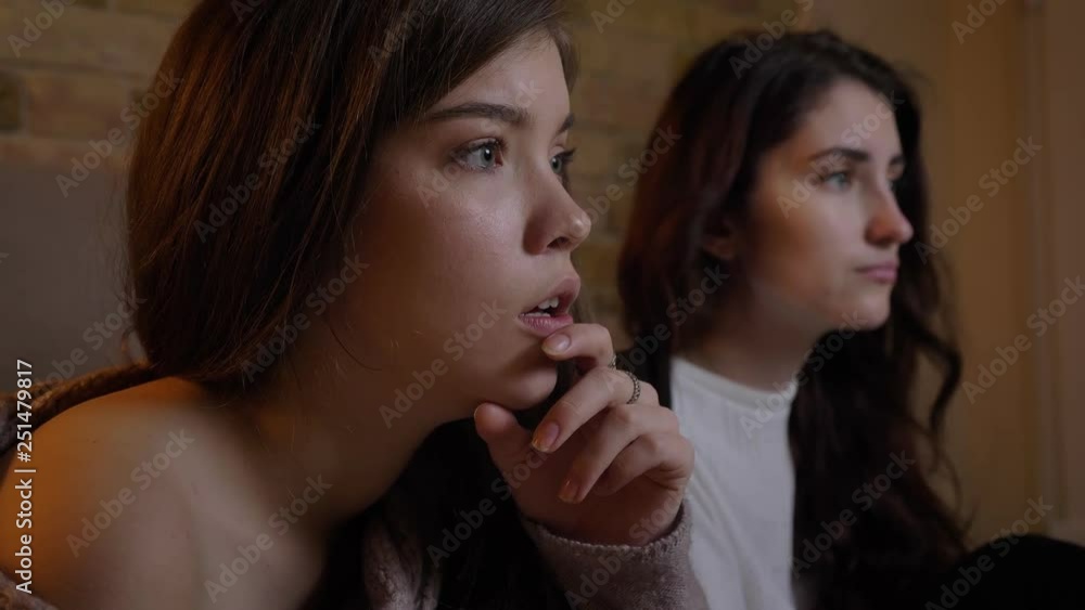 Close-up portrait in profile of scared young caucasian girls watching the TV seriously on cosy home background. Close-up portrait in profile of scared young caucasian girls watching the TV seriously on cosy home background.