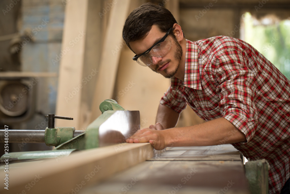 Confident and serious joiner grinding timber with help of special equipment. Busy carpenter in checked shirt and safety glasses working in joiner's shop. Handsome man working with wood. Confident and serious joiner grinding timber with help of special equipment. Busy carpenter in checked shirt and safety glasses working in joiner's shop. Handsome man working with wood.