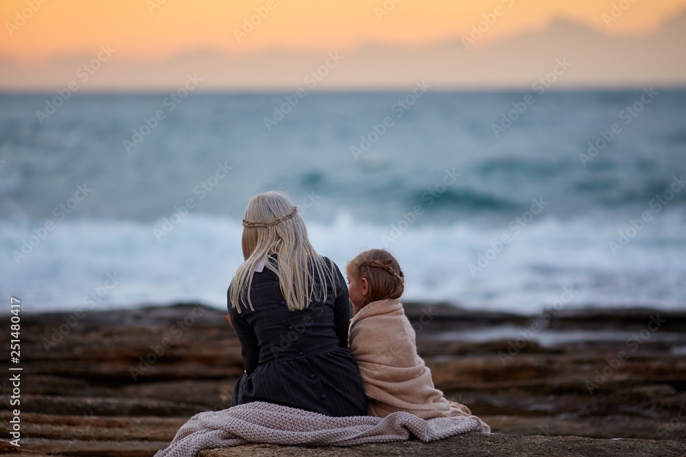 Back view of blond woman with little girl cuddling in plaid on rocky shoreline in twilight time