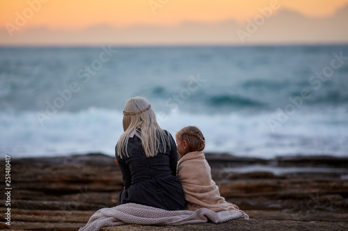 Back view of blond woman with little girl cuddling in plaid on rocky shoreline in twilight time