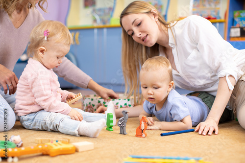 Nursery babies with teachers playing with wooden educational toys sitting on a floor indoor