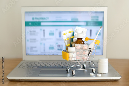 Shopping cart toy with medicaments in front of laptop screen with pharmacy web site on it. Pills, blister packs, medical bottles, thermometer set. Health care and internet shopping.