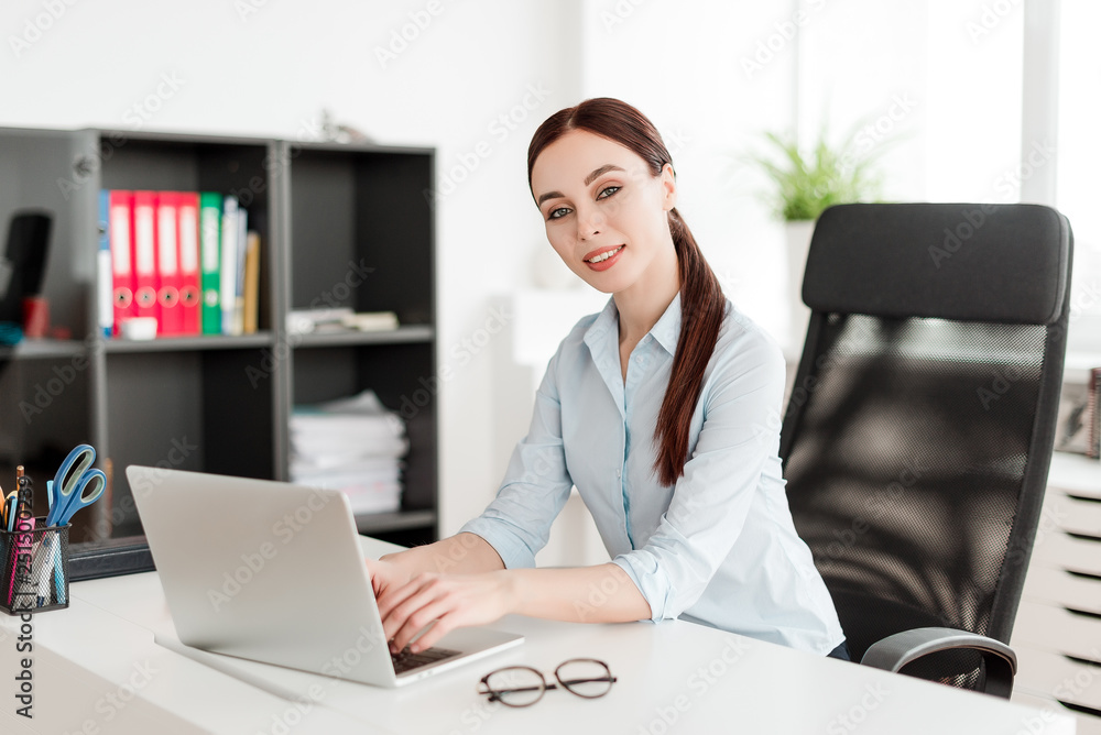 Business concept - successful young business woman smiling and sitting at her desk with the laptop in the office Business concept - successful young business woman smiling and sitting at her desk with the laptop in the office