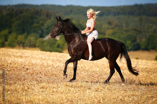 Horse with rider (woman) on a summer evening gallops over a harvested cornfield, wearing only a summer dress and bareback with open hair.