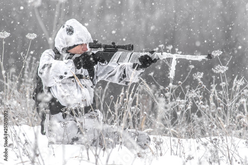 Man soldier in the winter on a hunt with a sniper rifle in white winter camouflage aims sitting in the snow