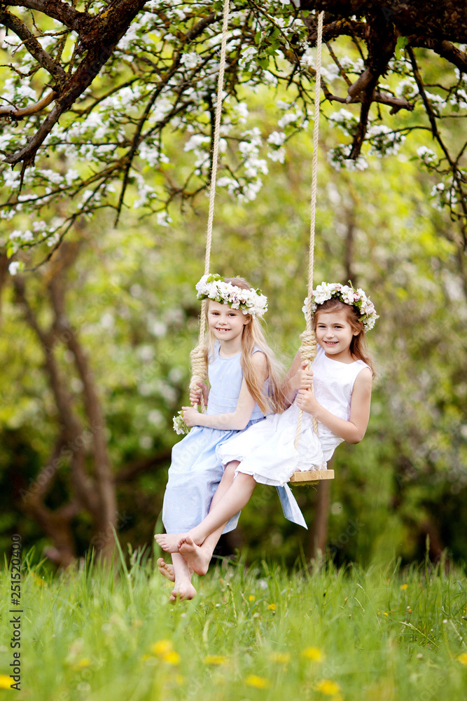 Two cute girls having fun on a swing in blossoming old apple tree garden. Sunny day. Spring outdoor activities for kids