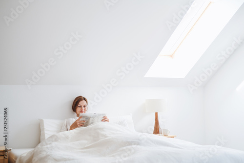 A young woman reading newspapers in bed in the morning in a bedroom.