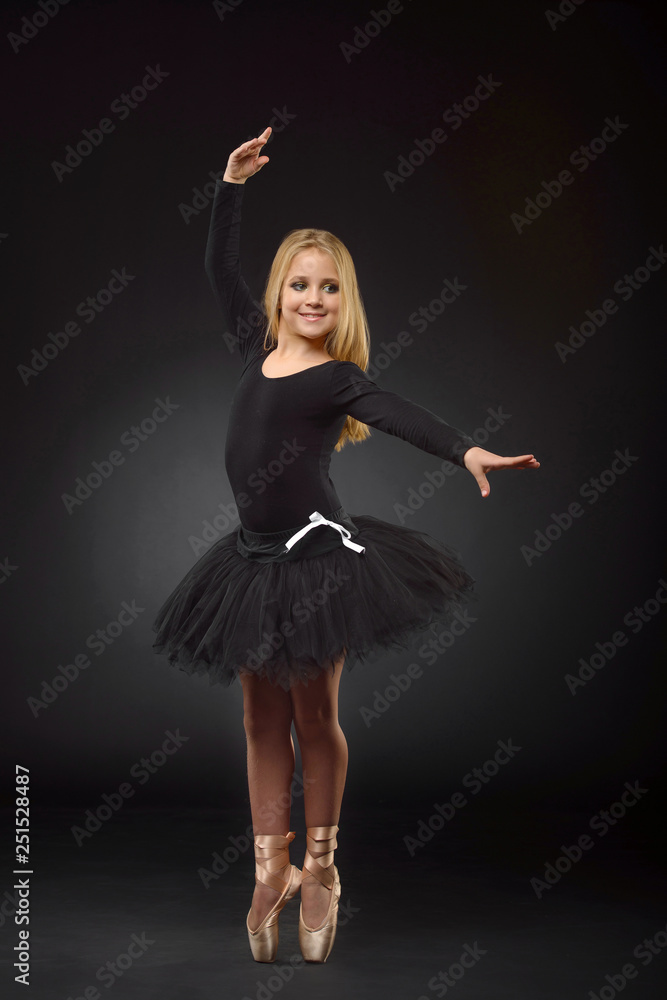 Cute little ballerina with long hair in a black tutu and pointe posing on a black background