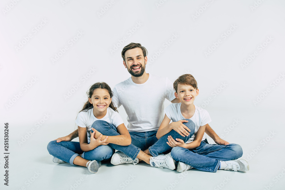 Handsome young father, attractive son and cute daughter are sitting together on the floor and hugging each other with happy and smiling faces. Best family Handsome young father, attractive son and cute daughter are sitting together on the floor and hugging each other with happy and smiling faces. Best family