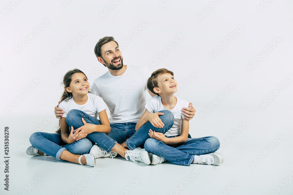 Handsome young father, attractive son and cute daughter are sitting together on the floor and hugging each other with happy and smiling faces. Best family Handsome young father, attractive son and cute daughter are sitting together on the floor and hugging each other with happy and smiling faces. Best family