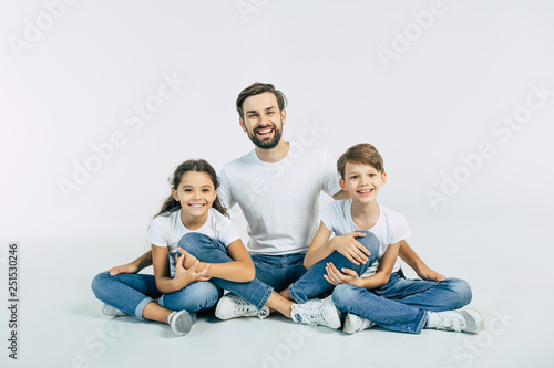 Handsome young father, attractive son and cute daughter are sitting together on the floor and hugging each other with happy and smiling faces. Best family