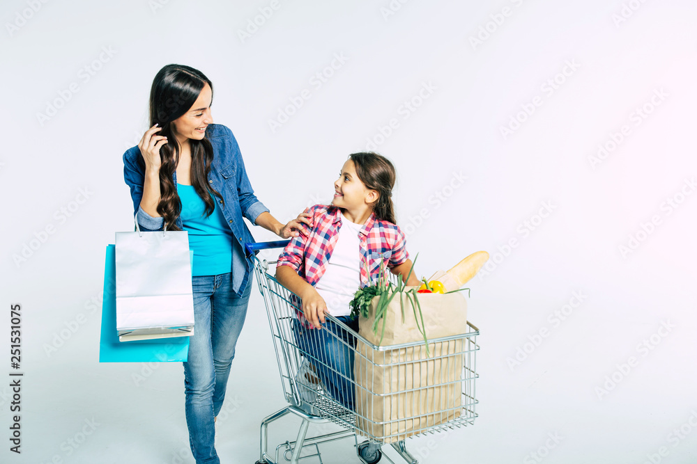 Attractive young mother with shopping bags is pushing her daughter who seat in food trolley with purchases and talking about something with smile. Attractive young mother with shopping bags is pushing her daughter who seat in food trolley with purchases and talking about something with smile.