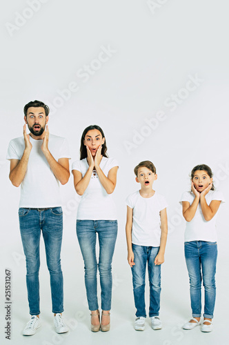 Shocked and surprised beautiful family in t-shirts standing in line looking on camera isolated on white background