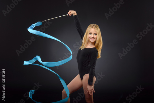 Young beautiful white caucasian girl gymnast doing gymnastic exercise with blue ribbon on black background.