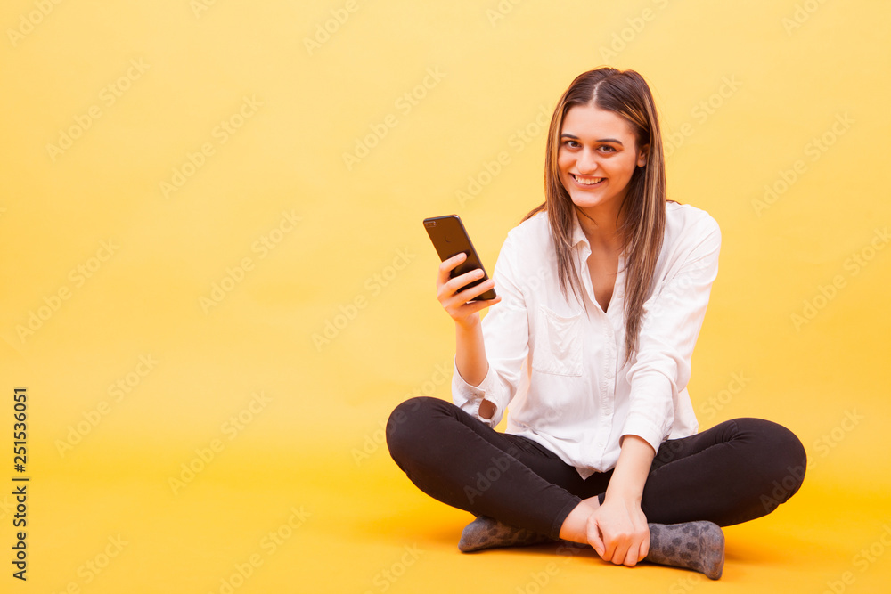 Beautiful girl smiling her phone while sitting down over yellow background Beautiful girl smiling her phone while sitting down over yellow background