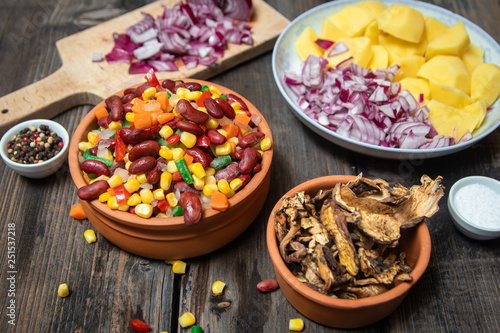 Mixture of vegetables, dried boletus mushrooms, sliced potato, red onion sliced and whole on a cutting board and spices, on old rustic wooden table.