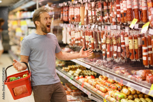 Client of supermarket shocked of prices on meat products. Bearded man standing with red basket in hand and looking at sausages and salami. Customer smiling with opened mouth.
