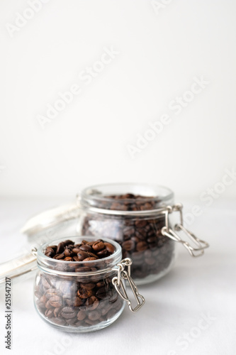 Dark roasted coffee beans in small and large glass jar on white cotton fabric background in natural light with empty text copy space on white wall background