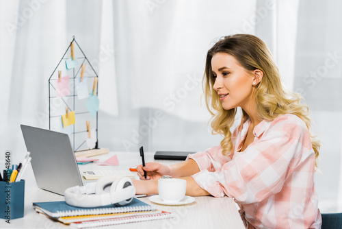 Fotografie Curious woman in checkered shirt writing in notebook and looking at laptop