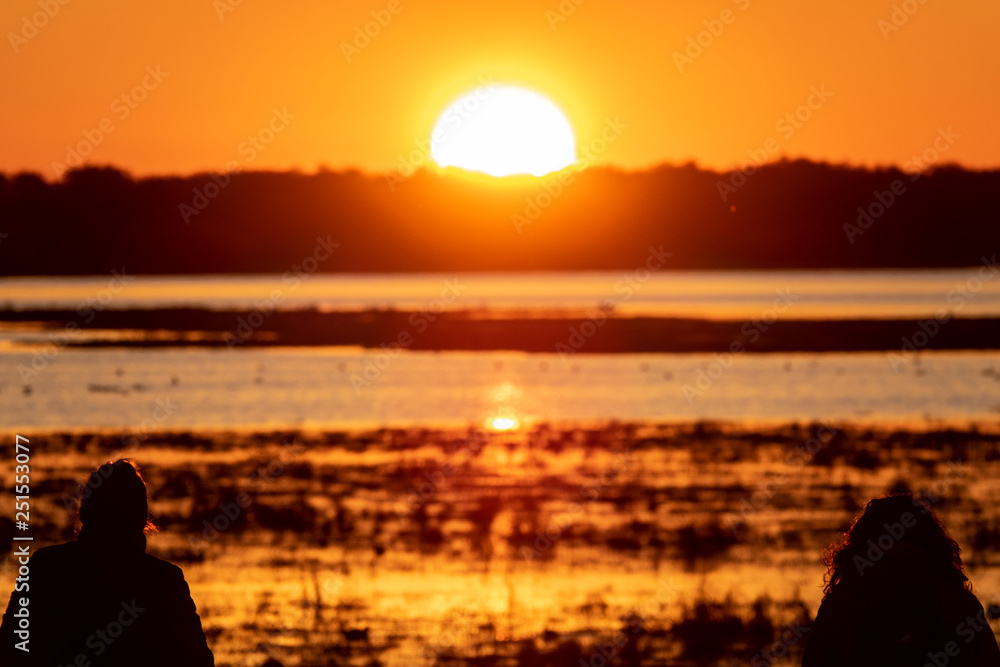 Two woman silhouettes observing sunrise over lake