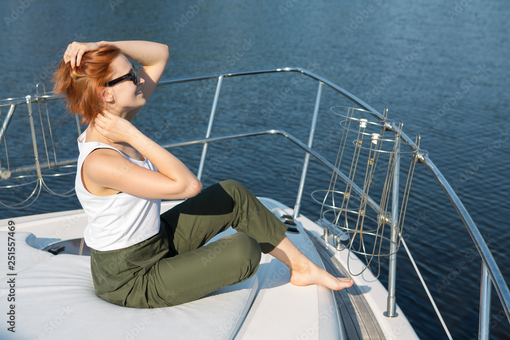 Sensuality and beautiful girl with short ginger hair posing at yacht, touching neck by hands and smiling. Pretty woman in black sunglasses sitting on boat bow and looking away in sea.