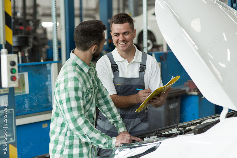 Two men standing in car service near white car and talking. Mechanic in uniform keeping papers and whiting contract and client in checkered shirt listening carefully. Concept of deal. Two men standing in car service near white car and talking. Mechanic in uniform keeping papers and whiting contract and client in checkered shirt listening carefully. Concept of deal.