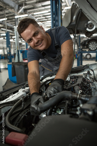 Front view of smiling worker of car service in process of repairing vehicle. Skilled man leaning over open hood, looking at camera and posing while fixing car. Concept of maintenance.