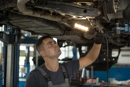 View from side of concentrated worker of car service checking running gear of automobile. Male mechanic in uniform and gloves standing under car, keeping lamp and looking for breakage.